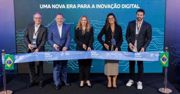 A imagem mostra uma cerimônia de inauguração ou abertura oficial, na qual cinco pessoas estão posicionadas lado a lado, cortando uma longa faixa azul. A faixa exibe os logotipos do BMW Group e do TechWorks Brasil, bem como textos institucionais relacionados às marcas. O fundo é composto por um painel grande em tom escuro, com elementos gráficos que remetem à tecnologia, como linhas e circuitos estilizados. Na parte superior do painel aparece a frase: “UMA NOVA ERA PARA A INOVAÇÃO DIGITAL” As pessoas presentes estão vestindo roupas formais ou semiformal corporativas, com crachás pendurados no pescoço. Cada uma segura uma tesoura, cortando simultaneamente a faixa cerimonial. À esquerda e à direita da cena há dois mastros com pequenas bandeiras do Brasil. O piso é um tapete escuro, e a iluminação é intensa e uniforme, destacando o painel ao fundo e a faixa sendo cortada.