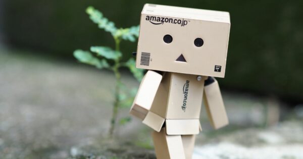 Uma pequena figura feita de caixas de papelão está posicionada ao ar livre, sobre uma superfície de concreto desfocada. A cabeça é uma caixa com o logotipo “amazon.co.jp” impresso na frente, e o rosto é composto por dois círculos pretos como olhos e um pequeno triângulo preto no centro como boca. O corpo e os membros também são formados por partes retangulares de papelão articuladas. A figura está levemente inclinada para o lado, como se estivesse dando um passo. Ao fundo, há um pequeno broto verde crescendo no solo e uma área desfocada em tons de verde e cinza. (Robos Amazon)