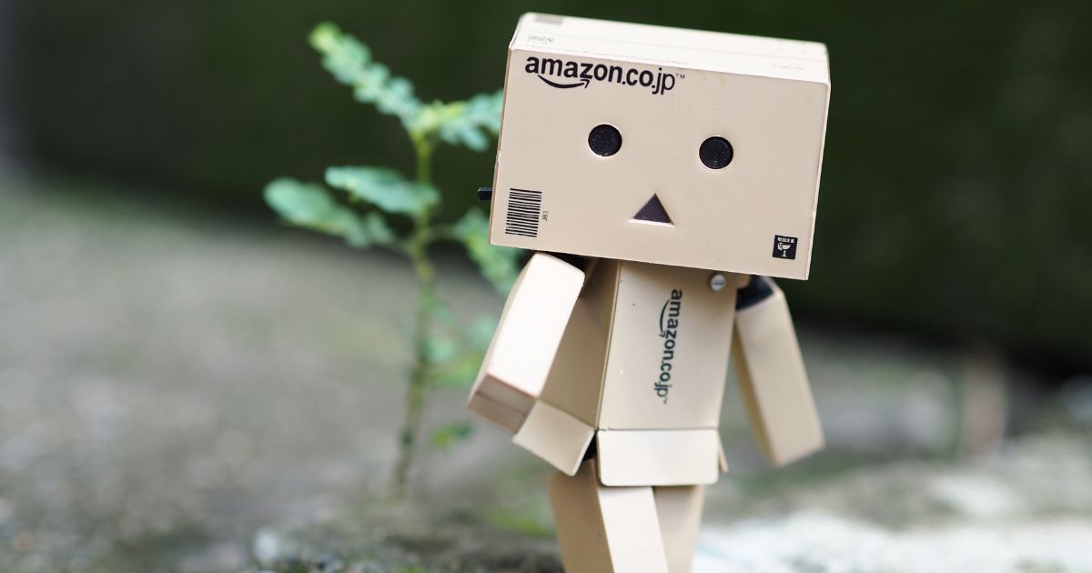Uma pequena figura feita de caixas de papelão está posicionada ao ar livre, sobre uma superfície de concreto desfocada. A cabeça é uma caixa com o logotipo “amazon.co.jp” impresso na frente, e o rosto é composto por dois círculos pretos como olhos e um pequeno triângulo preto no centro como boca. O corpo e os membros também são formados por partes retangulares de papelão articuladas. A figura está levemente inclinada para o lado, como se estivesse dando um passo. Ao fundo, há um pequeno broto verde crescendo no solo e uma área desfocada em tons de verde e cinza. (Robos Amazon)