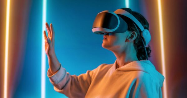 A imagem mostra um humano usando um headset de realidade virtual em um ambiente iluminado por luzes coloridas verticais em tons de azul, rosa e laranja. A pessoa está com um braço levantado e a mão estendida, como se estivesse interagindo com elementos digitais no espaço virtual. A iluminação cria reflexos suaves na roupa clara e enfatiza o clima futurista da cena, transmitindo a ideia de imersão em tecnologia de realidade virtual.