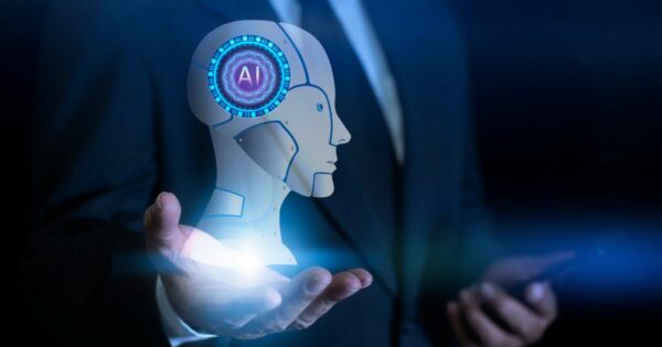 A imagem mostra uma pessoa usando traje formal escuro, segurando uma projeção holográfica em formato de cabeça robótica estilizada. O holograma apresenta detalhes futuristas, com anéis luminosos em azul ao redor de um ícone com as letras “AI” no centro. A mão está posicionada abaixo da projeção, como se estivesse sustentando o elemento digital. O fundo é escuro, com luzes suaves que reforçam a atmosfera tecnológica, e a outra mão segura um dispositivo semelhante a um smartphone. (inteligencia artificial)
