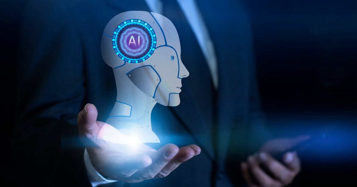 A imagem mostra uma pessoa usando traje formal escuro, segurando uma projeção holográfica em formato de cabeça robótica estilizada. O holograma apresenta detalhes futuristas, com anéis luminosos em azul ao redor de um ícone com as letras “AI” no centro. A mão está posicionada abaixo da projeção, como se estivesse sustentando o elemento digital. O fundo é escuro, com luzes suaves que reforçam a atmosfera tecnológica, e a outra mão segura um dispositivo semelhante a um smartphone. (inteligencia artificial)