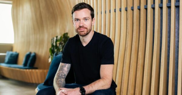 A imagem mostra uma pessoa sentada em um ambiente interno com design moderno, caracterizado por painéis verticais de madeira curvada ao fundo. A pessoa está vestindo uma camiseta preta e jeans escuros, com um relógio no pulso e uma tatuagem visível em um dos braços. Atrás, há um banco contínuo com almofadas escuras e algumas plantas posicionadas ao lado, compondo um espaço aconchegante e contemporâneo (Stark Infra)