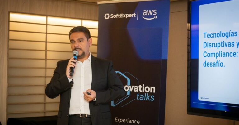 A imagem mostra uma pessoa vestida com traje social escuro, segurando um microfone com uma mão e um controle ou apontador com a outra, enquanto realiza uma apresentação em um evento corporativo. À esquerda, há um painel vertical com as marcas SoftExpert e AWS, acompanhado do título “Innovation Talks”. À direita, um telão exibe um slide com o texto: “Tecnologías Disruptivas y Compliance: desafío.” O ambiente tem iluminação quente e elementos visuais em azul, reforçando o clima profissional da apresentação.