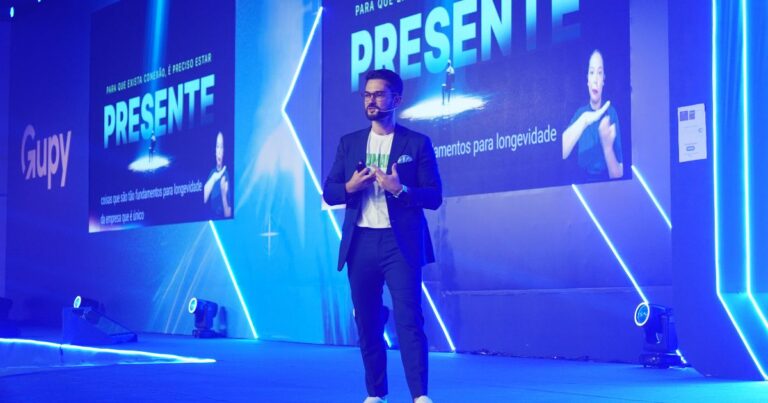 A imagem mostra uma pessoa no palco de um evento corporativo, iluminado por luzes azuis intensas e com painéis de LED ao fundo. Um grande telão exibe a palavra “PRESENTE” junto a elementos gráficos e texto adicional. A pessoa veste um traje casual elegante composto por blazer escuro, camiseta clara e tênis. O cenário inclui refletores no chão, marcações de palco e o logotipo Gupy projetado na lateral esquerda. O ambiente transmite a atmosfera de uma apresentação profissional em conferência.