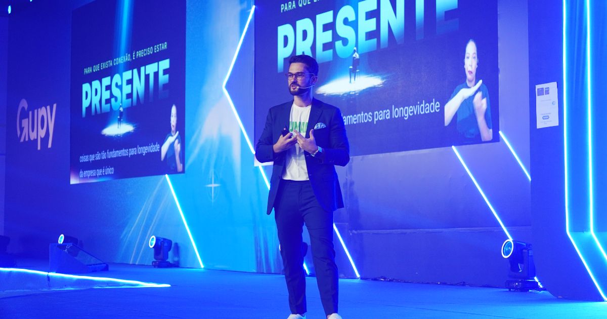 A imagem mostra uma pessoa no palco de um evento corporativo, iluminado por luzes azuis intensas e com painéis de LED ao fundo. Um grande telão exibe a palavra “PRESENTE” junto a elementos gráficos e texto adicional. A pessoa veste um traje casual elegante composto por blazer escuro, camiseta clara e tênis. O cenário inclui refletores no chão, marcações de palco e o logotipo Gupy projetado na lateral esquerda. O ambiente transmite a atmosfera de uma apresentação profissional em conferência.