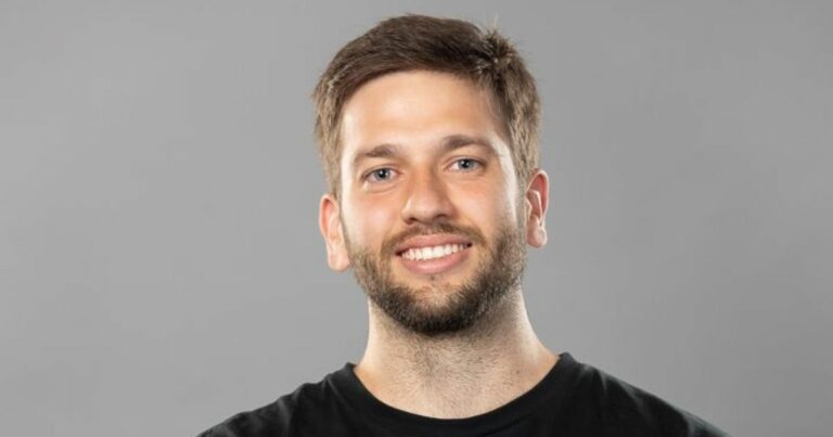 A imagem mostra uma pessoa em frente a um fundo neutro cinza, com iluminação suave e uniforme típica de estúdio. A pessoa está vestindo uma camiseta preta simples, e o enquadramento captura a parte superior do tronco. O cabelo é curto e bem aparado. A composição é minimalista, com foco no contraste entre a roupa escura e o fundo claro. (Rodrigo Stefanini)