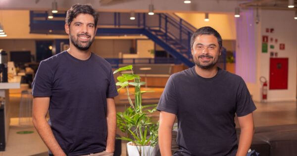 Duas pessoas estão em um ambiente interno moderno, que parece ser um escritório ou espaço de convivência corporativo. As duas estão posicionadas próximas a uma planta em vaso colocada sobre uma mesa baixa; uma delas está de pé à esquerda, enquanto a outra está sentada à direita. Ambas vestem camisetas escuras e calças. Ao fundo, há uma área ampla com iluminação quente, mesas, cadeiras e uma escada metálica azul que leva ao mezanino. À direita, na parede, encontram-se uma porta vermelha e alguns itens de sinalização. A iluminação do espaço é composta por diversas luminárias no teto. (Mogno, Accountfy)