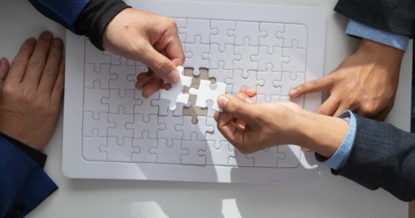 Três pessoas participam da montagem de um quebra‑cabeça branco sobre uma mesa. Duas mãos seguram uma peça central que está prestes a completar o espaço vazio no tabuleiro, enquanto outra mão repousa próxima ao canto. Todas as peças são brancas, criando um visual minimalista e organizado. (venture capital)