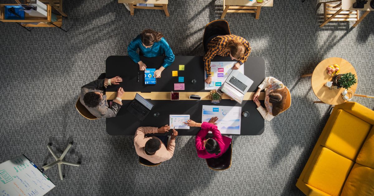 Imagem aérea de um grupo de seis pessoas reunidas ao redor de uma mesa retangular preta em um ambiente de escritório colaborativo. Sobre a mesa há laptops, tablets, cadernos, gráficos impressos, canecas, post-its coloridos e materiais de trabalho. As pessoas estão distribuídas em cadeiras ao redor da mesa, interagindo entre si e analisando documentos. O espaço ao redor inclui uma área com carpete cinza, uma poltrona amarela e pequenas mesas laterais com objetos decorativos. (venture)