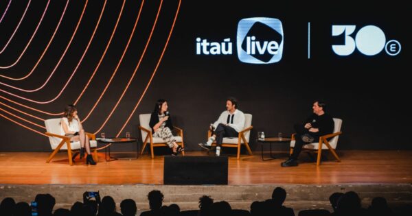 Uma imagem mostra um palco de auditório onde quatro pessoas estão sentadas em poltronas claras, dispostas em semicírculo, participando de uma conversa ou painel. Todas seguram microfones. Há duas mesas de centro pequenas entre elas, cada uma com copos de água. O fundo do palco é uma tela grande, predominantemente preta, exibindo elementos gráficos curvos em tons de laranja e rosa à esquerda, e os logotipos “itaú live” e “30e” à direita. Na frente do palco é possível ver silhuetas da plateia assistindo ao evento.