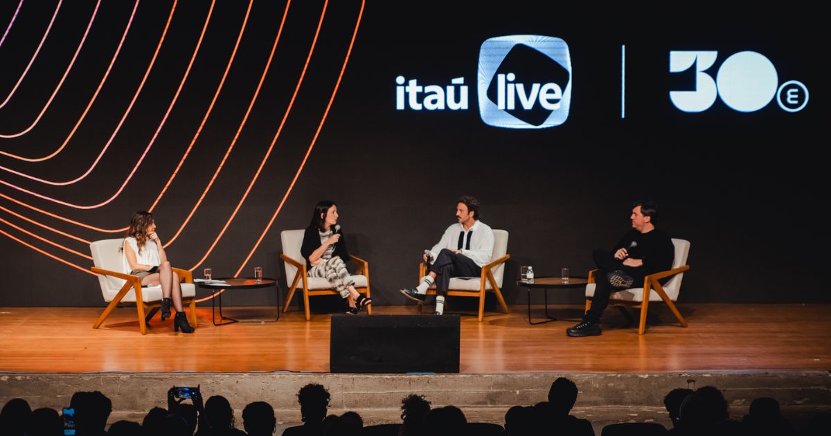 Uma imagem mostra um palco de auditório onde quatro pessoas estão sentadas em poltronas claras, dispostas em semicírculo, participando de uma conversa ou painel. Todas seguram microfones. Há duas mesas de centro pequenas entre elas, cada uma com copos de água. O fundo do palco é uma tela grande, predominantemente preta, exibindo elementos gráficos curvos em tons de laranja e rosa à esquerda, e os logotipos “itaú live” e “30e” à direita. Na frente do palco é possível ver silhuetas da plateia assistindo ao evento.
