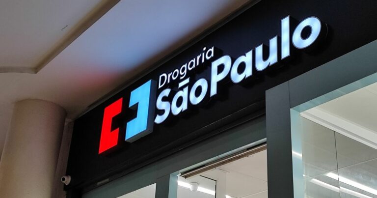 A imagem mostra a fachada de uma loja da Drogaria São Paulo vista de um ângulo levemente inclinado para cima. O letreiro luminoso tem as palavras “Drogaria São Paulo” escritas em letras brancas e brilhantes. À esquerda do texto há um símbolo composto por dois quadrados: um vermelho e outro azul‑claro, formando um design geométrico. A fachada é preta e, logo abaixo do letreiro, é possível ver parte da entrada da loja, com portas de vidro e iluminação interna branca. (Grupo DPSP)