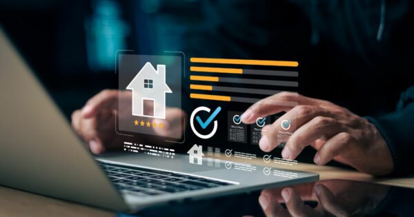 A imagem mostra uma pessoa utilizando um laptop em uma mesa, enquanto elementos gráficos digitais aparecem projetados acima do teclado. Entre os ícones visíveis há a silhueta de uma casa com cinco estrelas abaixo, um grande símbolo de verificação dentro de um círculo e diversos blocos de informações em formato de cartões. Barras horizontais coloridas, caixas de seleção marcadas e pequenos indicadores visuais compõem a interface. O ambiente é escuro, iluminado principalmente pelo brilho azulado da projeção digital e da tela do laptop, reforçando um clima tecnológico e moderno. (serviço)