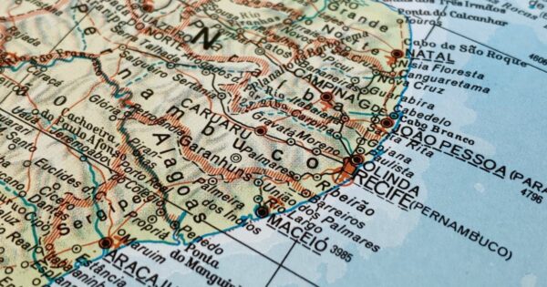 A imagem mostra um recorte de um mapa físico e político da região Nordeste do Brasil. Estão visíveis estados como Pernambuco, Paraíba, Rio Grande do Norte, Ceará, Alagoas e Sergipe, com seus limites estaduais demarcados. Diversas cidades aparecem nomeadas, incluindo Recife, Olinda, João Pessoa, Natal, Maceió, Aracaju, Caruaru, entre outras. A parte direita do mapa exibe o litoral voltado para o oceano, em tons azul‑claro que representam o mar. O interior dos estados está marcado por linhas, relevo e pontos que indicam rodovias, rios e localizações urbanas. As cores predominantes são bege, azul e vermelho, típicas de mapas impressos tradicionais.