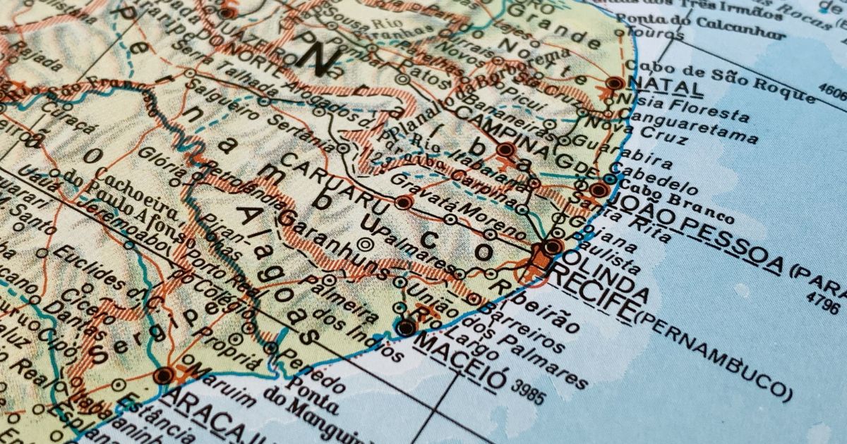 A imagem mostra um recorte de um mapa físico e político da região Nordeste do Brasil. Estão visíveis estados como Pernambuco, Paraíba, Rio Grande do Norte, Ceará, Alagoas e Sergipe, com seus limites estaduais demarcados. Diversas cidades aparecem nomeadas, incluindo Recife, Olinda, João Pessoa, Natal, Maceió, Aracaju, Caruaru, entre outras. A parte direita do mapa exibe o litoral voltado para o oceano, em tons azul‑claro que representam o mar. O interior dos estados está marcado por linhas, relevo e pontos que indicam rodovias, rios e localizações urbanas. As cores predominantes são bege, azul e vermelho, típicas de mapas impressos tradicionais.