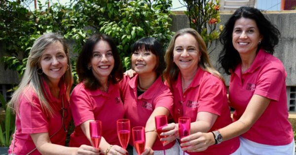 Um grupo de cinco mulheres está reunido ao ar livre, posicionadas lado a lado e inclinadas levemente para frente, segurando taças rosadas translúcidas em um gesto de brinde. Todas vestem camisetas polo na cor rosa e calças brancas. Ao fundo, há vegetação com arbustos verdes e, mais acima, estruturas de concreto e grades metálicas. A luz do sol é intensa, sugerindo um dia claro.