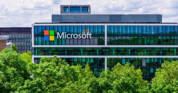 A imagem mostra um prédio corporativo moderno, com fachada de vidro refletivo em tons de verde e azul. No centro superior do edifício há o logotipo da Microsoft, composto pelo ícone quadrado dividido em quatro cores (vermelho, verde, azul e amarelo) seguido do nome “Microsoft” em letras brancas. Em primeiro plano, há uma copa de árvores densas, de folhagem verde vibrante, parcialmente ocultando a base do prédio. Ao fundo, o céu está parcialmente nublado, com nuvens brancas e luz suave, conferindo um aspecto claro e arejado à cena. Outros prédios contemporâneos aparecem discretamente ao lado e atrás da construção principal, sugerindo um ambiente urbano corporativo.