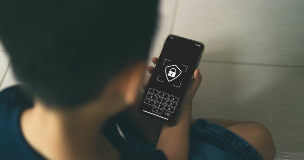 Uma criança sentada no chão segura um smartphone cuja tela exibe um cadeado e um teclado numérico de senha, sugerindo uma interface de bloqueio ou segurança digital. A cena é iluminada suavemente e o foco está no dispositivo.