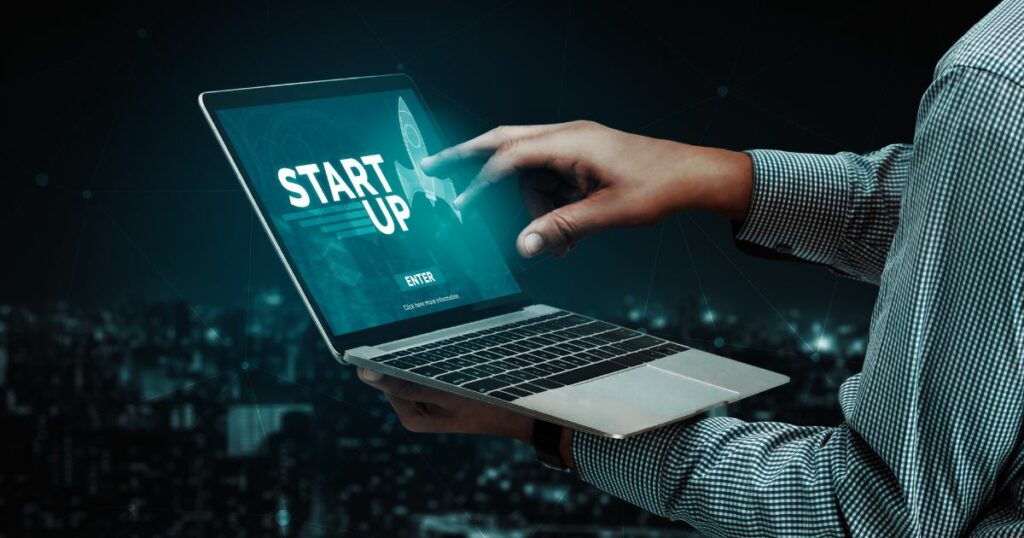 Pessoa segurando um laptop aberto enquanto toca a tela com uma das mãos. No display, aparece a palavra ‘STARTUP’ acompanhada da imagem de um foguete estilizado e um botão de ‘Enter’. Ao fundo, observa‑se uma cidade desfocada à noite, com diversas luzes urbanas criando um clima tecnológico e futurista. (sellentt, datamint)