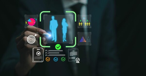 Imagem conceitual em ambiente escuro e corporativo mostrando uma mão humana interagindo com uma interface digital holográfica. No centro da interface, aparecem duas silhuetas humanas estilizadas, uma masculina e uma feminina, destacadas por um contorno verde com um ícone de verificação abaixo. Ao redor, há vários ícones gráficos representando dados e avaliações, como gráfico de pizza, alvo com setaó, lista de verificação, agrupamento de pessoas e pirâmide de níveis. A composição sugere análise de perfis, seleção de talentos, gestão de pessoas ou tomada de decisão baseada em dados. (IBSD)