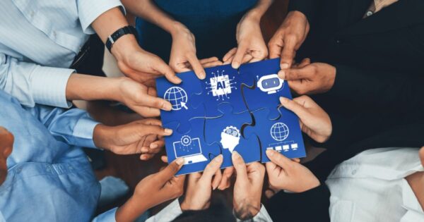 Imagem vista de cima mostrando várias mãos de diferentes pessoas unidas ao redor de uma peça central formada por partes de um quebra-cabeça azul. As mãos seguram as peças conectadas, formando um quadrado. Sobre as peças do quebra-cabeça aparecem ícones brancos relacionados à tecnologia e inovação, como um globo com lupa, um chip com a sigla “AI”, um robô, engrenagens, uma cabeça humana com circuitos e gráficos. O enquadramento enfatiza colaboração, trabalho em equipe e integração de ideias em torno de tecnologia e inteligência artificial. (dados)