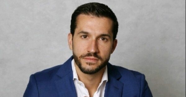 Retrato de uma pessoa posicionada de frente para a câmera, em ambiente interno com fundo liso e neutro em tom claro. A pessoa veste blazer azul sobre camisa clara com colarinho aberto. O enquadramento mostra o rosto e parte superior do tronco. O cabelo é curto e bem alinhado, e há barba curta. A iluminação é uniforme, destacando o rosto sem sombras marcadas, com fundo sem elementos visuais adicionais. (Simpress)