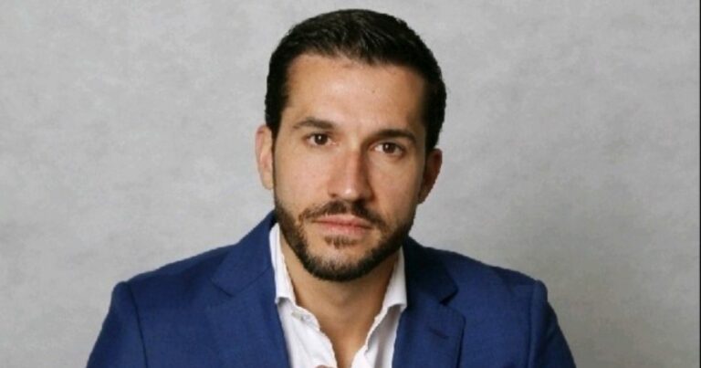 Retrato de uma pessoa posicionada de frente para a câmera, em ambiente interno com fundo liso e neutro em tom claro. A pessoa veste blazer azul sobre camisa clara com colarinho aberto. O enquadramento mostra o rosto e parte superior do tronco. O cabelo é curto e bem alinhado, e há barba curta. A iluminação é uniforme, destacando o rosto sem sombras marcadas, com fundo sem elementos visuais adicionais. (Simpress)
