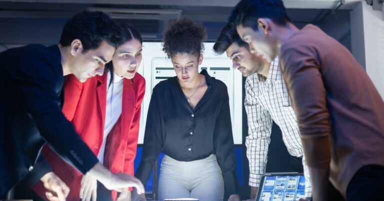 A imagem mostra um grupo de quatro pessoas reunidas ao redor de uma mesa em um ambiente interno com iluminação artificial. Todas estão inclinadas para a frente, observando algo sobre a mesa — possivelmente um dispositivo ou projeto iluminado. Uma tela com várias janelas ou gráficos está posicionada à direita. A cena transmite a ideia de colaboração em um trabalho técnico ou discussão de um projeto, com cada pessoa posicionada de forma concentrada e próxima ao centro da mesa. (liderança)