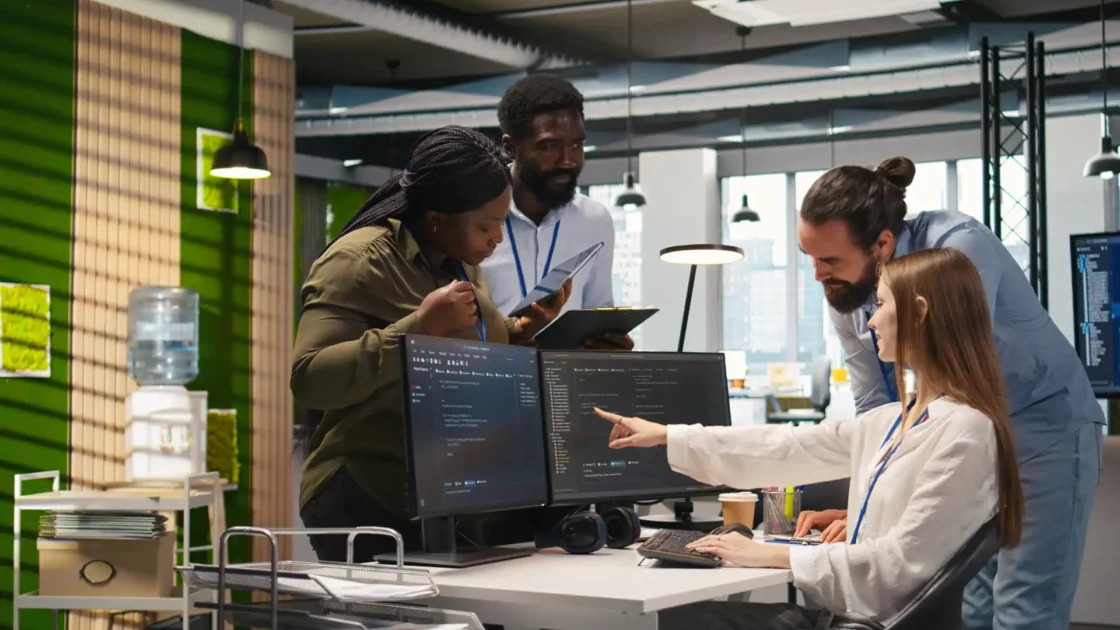 Inteligência emocional e autoconhecimento lideram buscas por soft skills no Brasil 2 Equipe de profissionais de tecnologia discutindo projeto de software em ambiente de escritório moderno, com dois computadores e profissionais colaborando. (soft Skills, autoconhecimento)