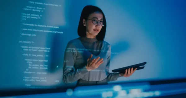 Mulher jovem usando óculos e segurando um tablet, trabalhando com códigos de programação em um ambiente com luz azul e tecnologia avançada.
