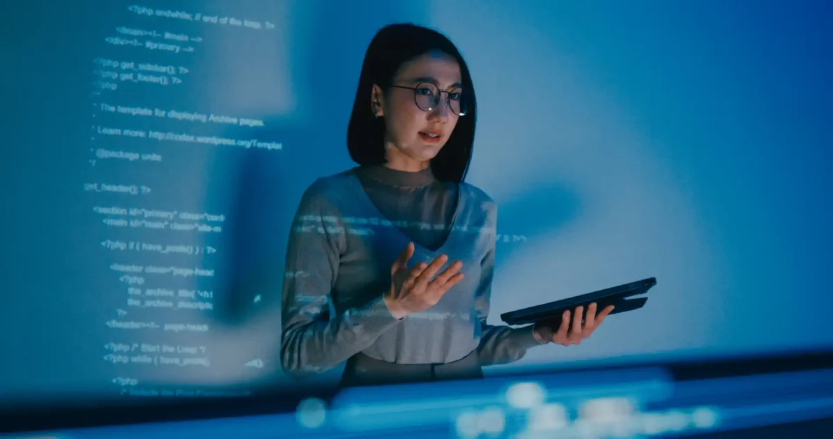 Mulher jovem usando óculos e segurando um tablet, trabalhando com códigos de programação em um ambiente com luz azul e tecnologia avançada.