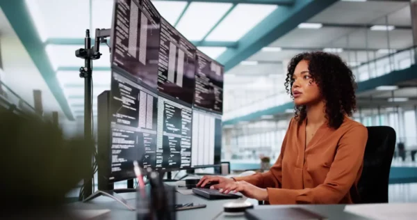 Mulher trabalhando em escritório moderno com múltiplos monitores exibindo códigos de programação, foco e concentração, ambiente de alta tecnologia. curso