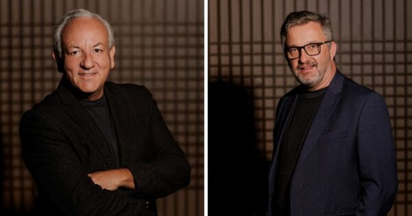 Imagem composta por dois retratos posicionados lado a lado, separados por uma faixa vertical clara. À esquerda, uma pessoa em pé, de frente para a câmera, com cabelos curtos grisalhos, vestindo blazer escuro sobre camiseta escura, com os braços cruzados diante do corpo. À direita, outra pessoa em pé, também de frente para a câmera, com cabelos curtos grisalhos, barba aparada e óculos, vestindo blazer azul escuro sobre camiseta escura. Ambas estão em ambiente interno com fundo padronizado em tons de marrom, formando uma grade geométrica. A iluminação é controlada e direcionada, destacando as pessoas em primeiro plano e criando um retrato de aparência profissional. (agentes)