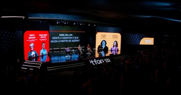 Imagem panorâmica de um grande palco em um evento corporativo ou tecnológico, visto a partir da plateia. O palco é amplo, com piso escuro e iluminação cênica profissional. Na parte frontal do palco, aparece o texto “itforum” em letras brancas grandes. Sobre o palco, várias pessoas estão sentadas em cadeiras individuais, participando de um painel de discussão, cada uma com uma pequena mesa lateral. Ao fundo, há grandes telas digitais coloridas. No painel central, lê-se o texto: “PAINEL CIOs: GENTE E AGENTES O QUE MUDA A PARTIR DE AGORA?”. Nas telas laterais, aparecem retratos estilizados de participantes acompanhados de nomes visíveis, como “RENATA MARQUES”, “EDUARDO GOMES”, “SUZANA RUBINIC” e “DÉBORAH OLIVEIRA”. À direita do palco, um painel circular exibe o logotipo “itforum”. A plateia aparece em primeiro plano, em área escura, com várias pessoas sentadas assistindo à apresentação. O conjunto transmite o contexto de uma conferência ou fórum profissional focado em tecnologia e inovação. (RH)