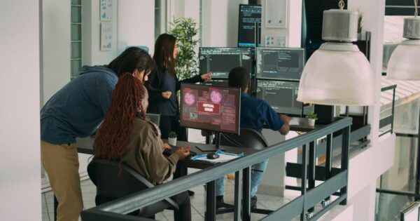 Imagem de um ambiente interno de escritório moderno e bem iluminado, com várias pessoas reunidas em torno de mesas de trabalho equipadas com múltiplos monitores. Em primeiro plano, duas pessoas estão sentadas diante de telas de computador; uma delas utiliza um teclado e mouse, enquanto a outra observa a tela. Em uma das telas centrais aparece um aviso em destaque com o texto “CRITICAL ERROR”, sobreposto a gráficos e códigos em tons de vermelho e preto. Outras pessoas estão em pé ao redor, observando os monitores e apontando para as telas, sugerindo análise conjunta do conteúdo exibido. Os monitores mostram linhas de código, interfaces técnicas e painéis de dados em fundo escuro. O ambiente conta com mesas pretas, cadeiras de escritório, luminárias grandes suspensas, plantas decorativas e paredes claras. Ao fundo, há janelas amplas que permitem a entrada de luz natural, reforçando o aspecto contemporâneo e tecnológico do espaço.