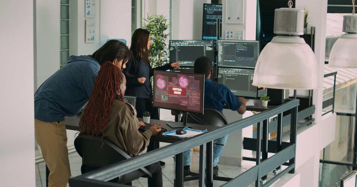 Imagem de um ambiente interno de escritório moderno e bem iluminado, com várias pessoas reunidas em torno de mesas de trabalho equipadas com múltiplos monitores. Em primeiro plano, duas pessoas estão sentadas diante de telas de computador; uma delas utiliza um teclado e mouse, enquanto a outra observa a tela. Em uma das telas centrais aparece um aviso em destaque com o texto “CRITICAL ERROR”, sobreposto a gráficos e códigos em tons de vermelho e preto. Outras pessoas estão em pé ao redor, observando os monitores e apontando para as telas, sugerindo análise conjunta do conteúdo exibido. Os monitores mostram linhas de código, interfaces técnicas e painéis de dados em fundo escuro. O ambiente conta com mesas pretas, cadeiras de escritório, luminárias grandes suspensas, plantas decorativas e paredes claras. Ao fundo, há janelas amplas que permitem a entrada de luz natural, reforçando o aspecto contemporâneo e tecnológico do espaço.