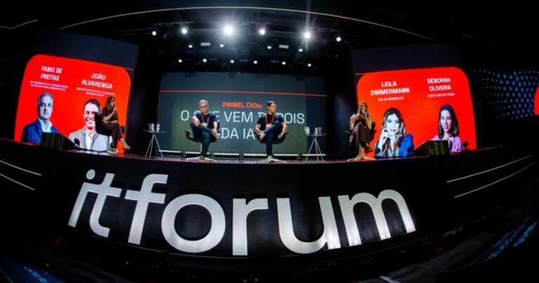Imagem de um grande palco em um evento corporativo ou tecnológico. Em primeiro plano, na borda frontal do palco, aparece o texto “itforum” em letras brancas grandes sobre fundo escuro. No palco, várias pessoas estão sentadas em cadeiras individuais, distribuídas ao longo do espaço, participando de um painel. Cada pessoa tem uma pequena mesa lateral com garrafas de água. Ao fundo, há grandes painéis digitais vermelhos e pretos. No painel central, lê‑se o texto “PAINEL CIOs: VEM DEPOIS DA IA?”. Nos painéis laterais, aparecem nomes e retratos estilizados de participantes, com textos visíveis como “FABIO DE FREITAS”, “JOÃO ALVARENGA”, “LEILA ZIMMERMANN” e “DÉBORAH OLIVEIRA”. A iluminação é profissional, com holofotes direcionados ao palco, criando um ambiente de conferência formal e tecnológico. (inteligência artificial)