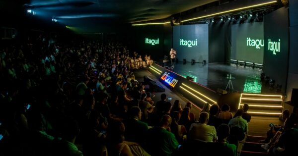 Imagem panorâmica de um grande auditório durante um evento corporativo ou tecnológico. Em primeiro plano e à esquerda, há uma plateia numerosa sentada em fileiras, ocupando quase todo o espaço do auditório, com iluminação baixa voltada ao público. À direita, um palco amplo e elevado é iluminado por luzes cênicas. No centro do palco, uma pessoa está em pé, falando ao público. Ao fundo do palco, grandes telas digitais exibem o texto “itaqui” em letras verdes sobre fundo escuro. Na borda frontal do palco, aparece o logotipo “it forum” iluminado. O palco possui degraus laterais com iluminação linear em tons claros. O ambiente é fechado, com teto escuro, estrutura de iluminação profissional e atmosfera de conferência formal voltada à tecnologia e inovação.