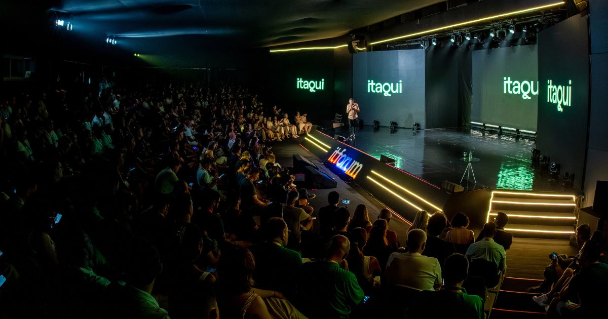 Imagem panorâmica de um grande auditório durante um evento corporativo ou tecnológico. Em primeiro plano e à esquerda, há uma plateia numerosa sentada em fileiras, ocupando quase todo o espaço do auditório, com iluminação baixa voltada ao público. À direita, um palco amplo e elevado é iluminado por luzes cênicas. No centro do palco, uma pessoa está em pé, falando ao público. Ao fundo do palco, grandes telas digitais exibem o texto “itaqui” em letras verdes sobre fundo escuro. Na borda frontal do palco, aparece o logotipo “it forum” iluminado. O palco possui degraus laterais com iluminação linear em tons claros. O ambiente é fechado, com teto escuro, estrutura de iluminação profissional e atmosfera de conferência formal voltada à tecnologia e inovação.