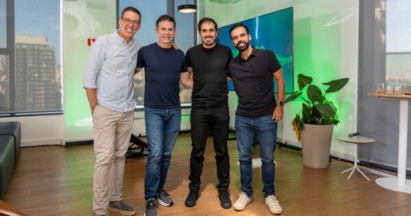 Imagem de quatro pessoas em pé, lado a lado, em um ambiente interno corporativo moderno. As quatro pessoas estão com os braços próximos ou apoiados umas nas outras, formando um grupo centralizado no enquadramento. Todas vestem roupas casuais em tons neutros, como camisetas e camisas de mangas longas combinadas com calças jeans ou claras. O ambiente tem piso de madeira e grandes janelas à esquerda, pelas quais é possível ver prédios ao fundo. À direita, há uma planta grande em vaso claro e uma pequena mesa lateral. Atrás do grupo, há uma tela grande ou painel iluminado em tons de verde e azul, sugerindo um espaço de escritório, sala de apresentação ou área de convivência corporativa. A iluminação é uniforme e bem distribuída. (Sankhya)