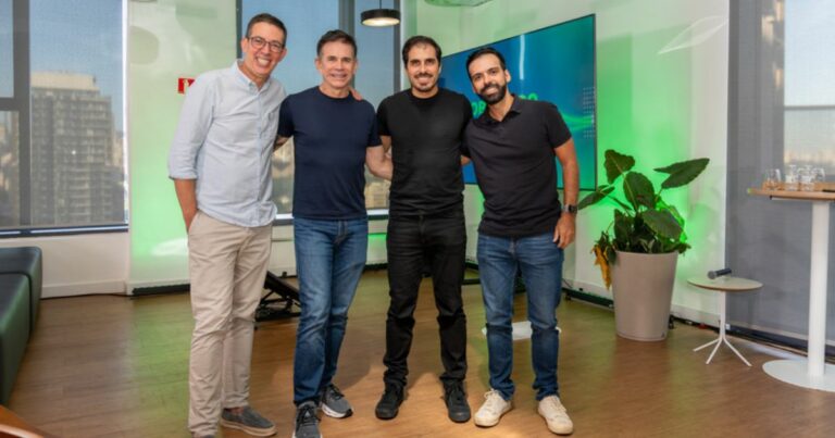 Imagem de quatro pessoas em pé, lado a lado, em um ambiente interno corporativo moderno. As quatro pessoas estão com os braços próximos ou apoiados umas nas outras, formando um grupo centralizado no enquadramento. Todas vestem roupas casuais em tons neutros, como camisetas e camisas de mangas longas combinadas com calças jeans ou claras. O ambiente tem piso de madeira e grandes janelas à esquerda, pelas quais é possível ver prédios ao fundo. À direita, há uma planta grande em vaso claro e uma pequena mesa lateral. Atrás do grupo, há uma tela grande ou painel iluminado em tons de verde e azul, sugerindo um espaço de escritório, sala de apresentação ou área de convivência corporativa. A iluminação é uniforme e bem distribuída. (Sankhya)