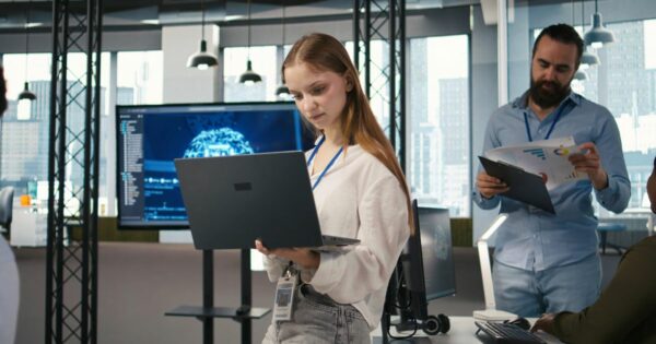 Imagem de um ambiente interno corporativo moderno, com várias pessoas utilizando equipamentos de tecnologia. Em primeiro plano, uma pessoa está em pé segurando um laptop aberto, vestindo roupa clara e usando um crachá no pescoço. Ao fundo, outra pessoa segura uma prancheta com documentos impressos. Há mesas de trabalho com computadores, teclados e fones de ouvido. Um monitor grande exibe uma interface digital com gráficos e elementos tecnológicos em tons de azul. O espaço possui grandes janelas com vista para prédios urbanos, estrutura metálica aparente no teto e iluminação uniforme, sugerindo um escritório de tecnologia ou inovação. (juniores)