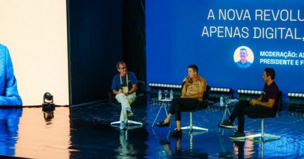 Imagem de um palco em ambiente interno durante um evento ou conferência. Quatro pessoas estão sentadas em cadeiras individuais, distribuídas em linha horizontal sobre um palco com piso escuro e reflexivo. Cada pessoa tem uma pequena mesa lateral com garrafa de água ao lado. Ao fundo, há um grande painel digital azul exibindo texto em português, parcialmente visível, com destaque para a frase “A NOVA REVOLUÇÃO … APENAS DIGITAL,” além de uma seção com o título “MODERAÇÃO:” seguida de texto menor e um ícone circular com a imagem de uma pessoa. À esquerda da imagem, parte de uma grande tela vertical clara também é visível. A iluminação é profissional, com luzes direcionadas ao palco, reforçando o contexto de um painel de discussão ou apresentação pública. (Semantix e Atos)