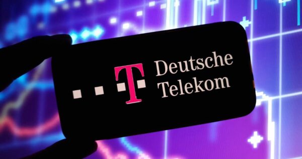 Imagem em close de um smartphone segurado na horizontal por uma mão em silhueta. Na tela do aparelho, com fundo preto, aparece o logotipo da Deutsche Telekom, composto por um “T” maiúsculo em magenta acompanhado de pequenos quadrados na mesma cor, seguido do texto “Deutsche Telekom” em letras claras. Ao fundo, desfocado, há gráficos financeiros ou de dados em tons de azul e roxo, com linhas, barras e símbolos luminosos que lembram visualizações de mercado ou tecnologia digital. O contraste entre a tela escura do celular e o fundo colorido destaca o logotipo exibido.