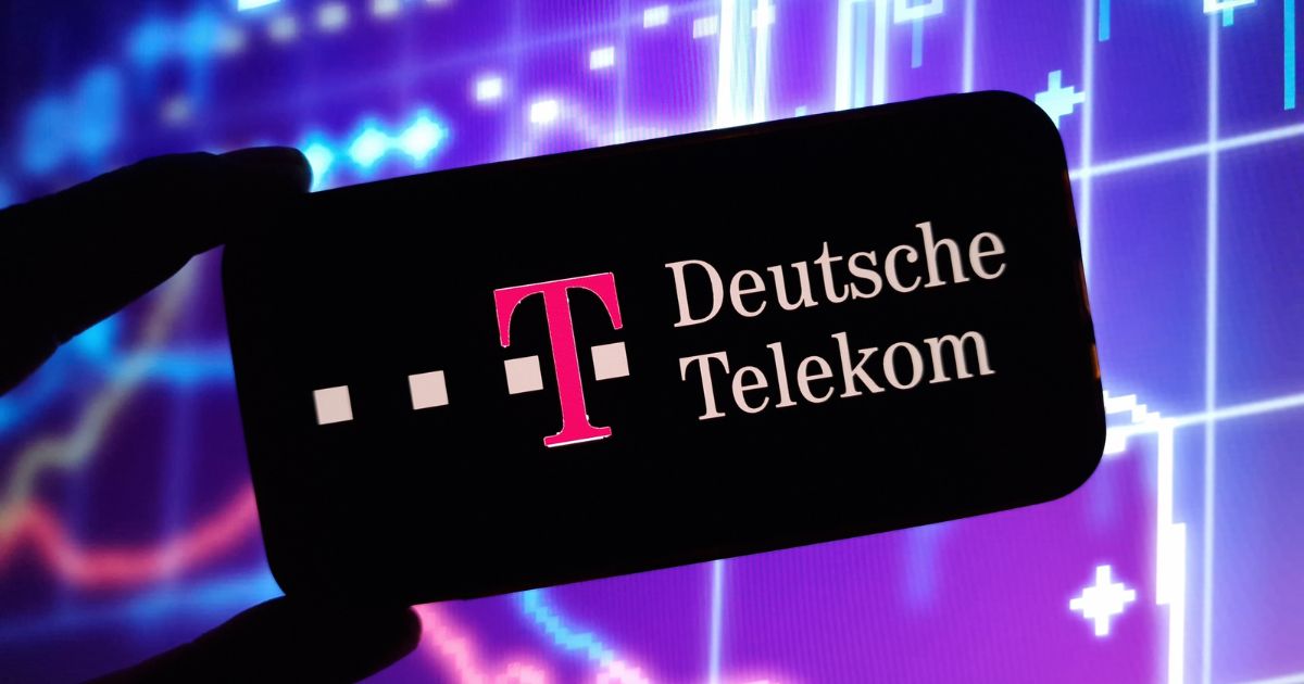 Imagem em close de um smartphone segurado na horizontal por uma mão em silhueta. Na tela do aparelho, com fundo preto, aparece o logotipo da Deutsche Telekom, composto por um “T” maiúsculo em magenta acompanhado de pequenos quadrados na mesma cor, seguido do texto “Deutsche Telekom” em letras claras. Ao fundo, desfocado, há gráficos financeiros ou de dados em tons de azul e roxo, com linhas, barras e símbolos luminosos que lembram visualizações de mercado ou tecnologia digital. O contraste entre a tela escura do celular e o fundo colorido destaca o logotipo exibido.