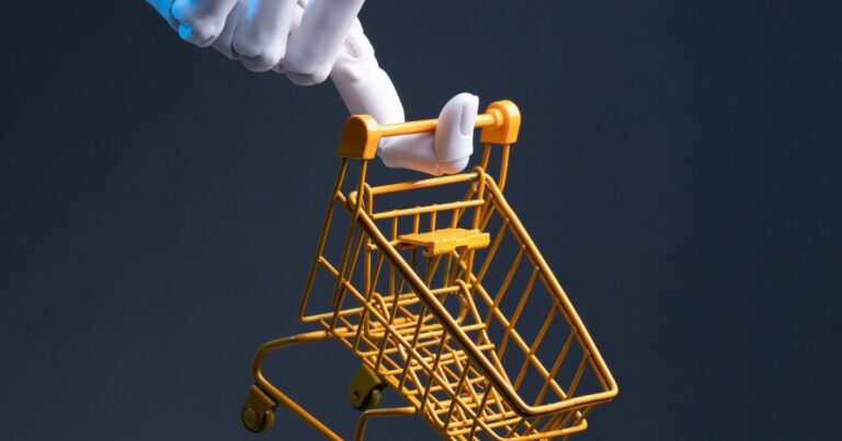 Imagem conceitual mostrando uma mão robótica branca e azul segurando a alça de um carrinho de compras em miniatura, de metal, na cor amarela. O carrinho está inclinado levemente para a frente, como se estivesse sendo empurrado. O fundo é liso e escuro, em tom azul‑acinzentado, sem outros elementos visíveis. A iluminação é direcionada, destacando a mão robótica e o carrinho, criando contraste e enfatizando a relação entre tecnologia e consumo ou varejo.