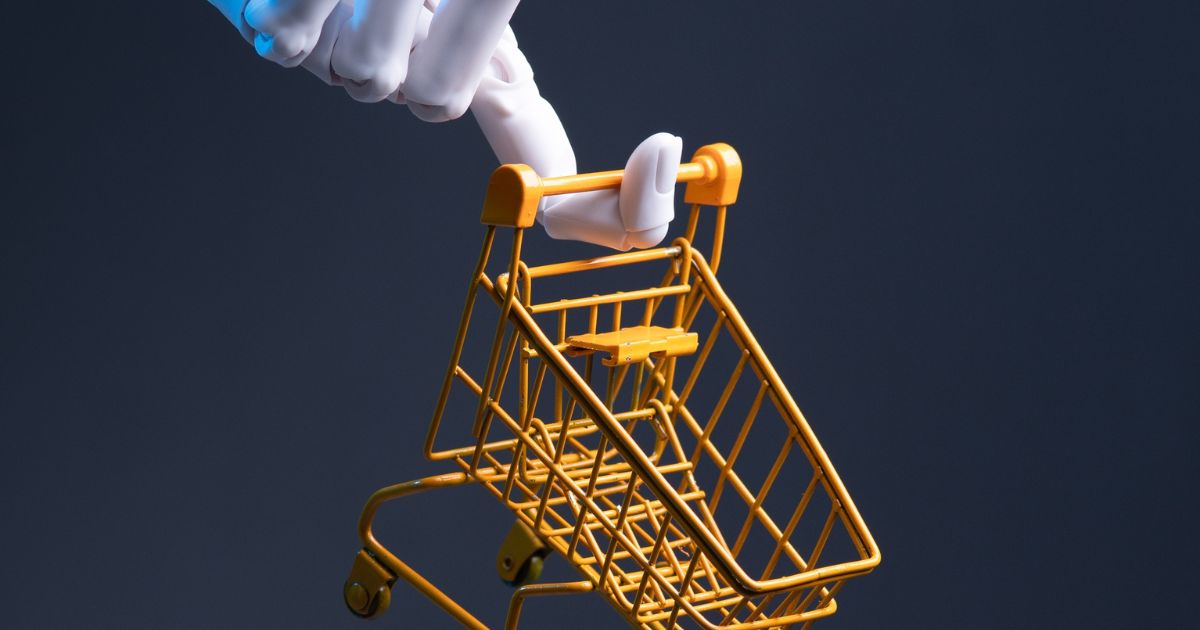 Imagem conceitual mostrando uma mão robótica branca e azul segurando a alça de um carrinho de compras em miniatura, de metal, na cor amarela. O carrinho está inclinado levemente para a frente, como se estivesse sendo empurrado. O fundo é liso e escuro, em tom azul‑acinzentado, sem outros elementos visíveis. A iluminação é direcionada, destacando a mão robótica e o carrinho, criando contraste e enfatizando a relação entre tecnologia e consumo ou varejo.
