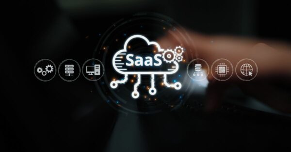 Imagem conceitual em fundo escuro mostrando um ícone luminoso de uma nuvem com a sigla “SaaS” no centro. A nuvem está conectada a pequenos pontos e linhas que se estendem para baixo, representando integração e distribuição de serviços. Ao redor da nuvem, aparecem ícones circulares em tom claro que simbolizam tecnologia e infraestrutura digital, como engrenagens, servidor, banco de dados, chip, globo e rede. Em primeiro plano, desfocado, observa‑se parte de uma mão se aproximando do centro da interface, sugerindo interação humana com uma plataforma de software como serviço. A composição transmite conceitos de computação em nuvem, automação e serviços digitais.