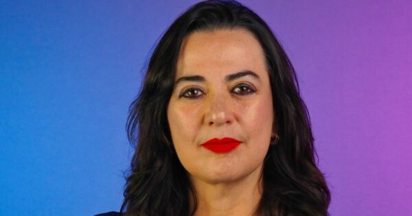 Retrato em close de uma pessoa posicionada de frente para a câmera. A pessoa tem cabelo escuro, liso e solto até abaixo dos ombros, e usa batom em tom vermelho intenso. O enquadramento mostra o rosto e parte superior do tronco. O fundo é liso com um degradê de cores, variando do azul à esquerda ao roxo à direita. A iluminação é uniforme e frontal, destacando os traços do rosto de maneira equilibrada, com aparência profissional e estúdio controlado. (Quatrix)